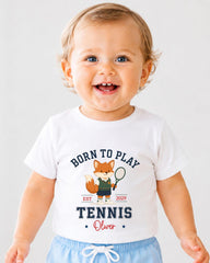 A smiling baby boy wearing a white t-shirt with a tennis-themed graphic and text.
