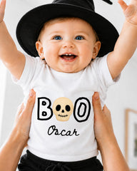 A smiling baby wearing a white t-shirt with a skull design and the text "BOO Oscar" being held up by someone's hands.
