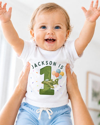 A smiling baby wearing a white t-shirt with the text "JACKSON IS 1" and a graphic of a green airplane with balloons.