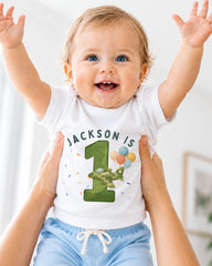 A smiling baby wearing a white t-shirt with the text "JACKSON IS 1" and a graphic of a green airplane with balloons.