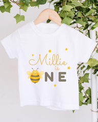 A white t-shirt with a yellow bee graphic and the text "Milly is one" printed on it, hanging on a wooden hanger.
