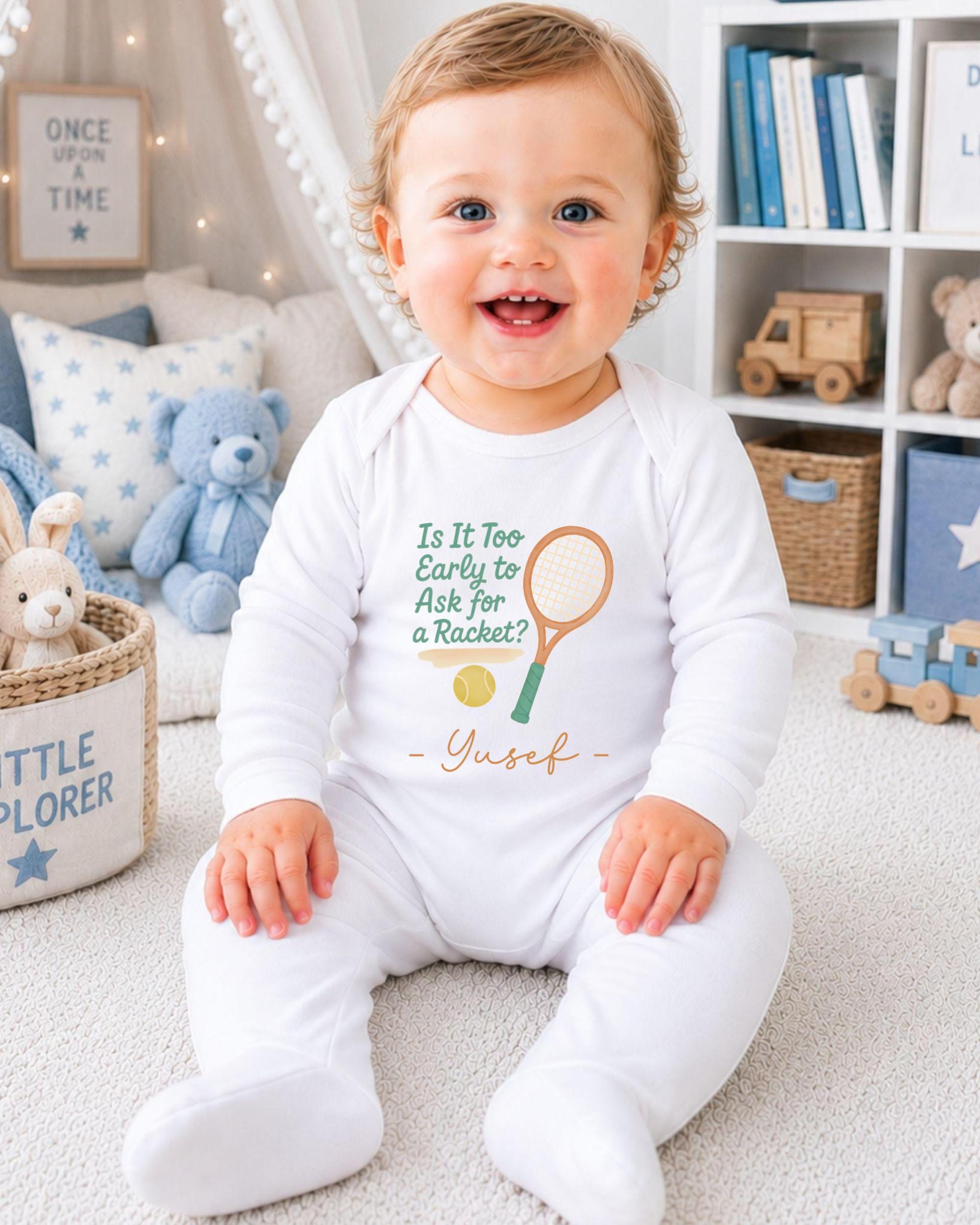 A smiling baby boy wearing a white onesie with a tennis racket and ball design, sitting on a carpeted floor in a nursery setting with bookshelves and stuffed animals in the background.