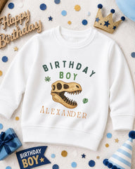 A white sweatshirt with a dinosaur skull design and the text "Birthday Boy Alexander" printed on it, surrounded by birthday decorations and a crown.
