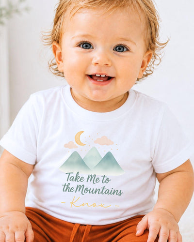 A smiling baby wearing a white t-shirt with a mountain and moon graphic and the text "Take Me to the Mountains - Knox" printed on it.