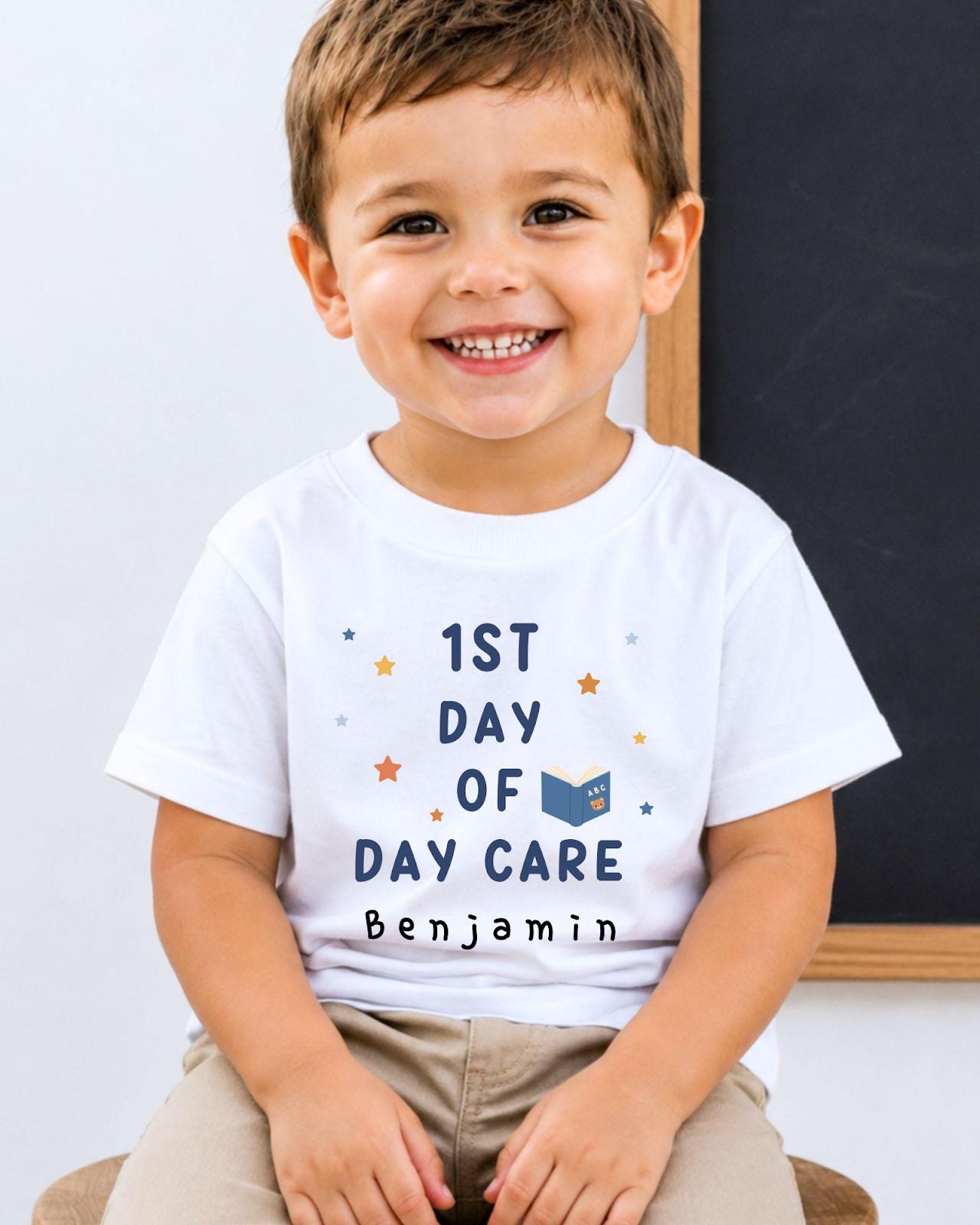 A young boy wearing a white t-shirt with the text "1st DAY OF DAY CARE Benjamin" is smiling at the camera.