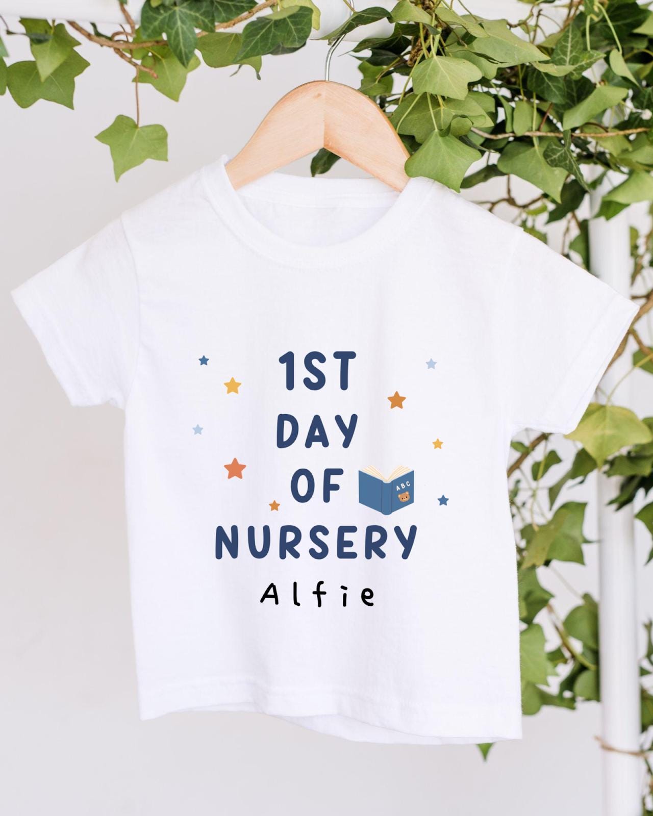 A white t-shirt with the text "1st DAY OF NURSERY" printed on it, along with a small illustration of a book and stars. The t-shirt is hanging on a wooden hanger, and there are green leaves visible in the background.