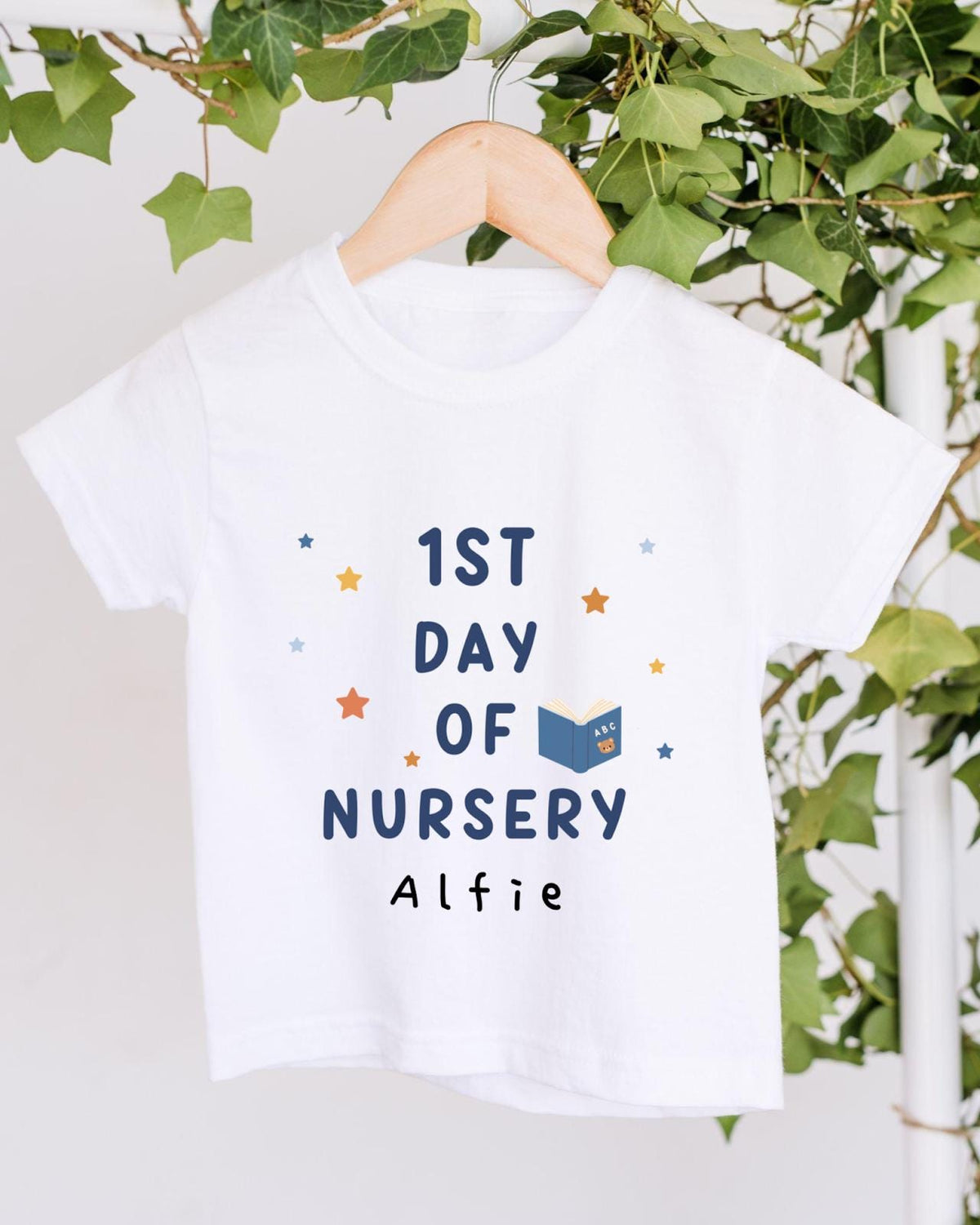 A white t-shirt with the text "1st DAY OF NURSERY" printed on it, along with a small illustration of a book and stars. The t-shirt is hanging on a wooden hanger, and there are green leaves visible in the background.