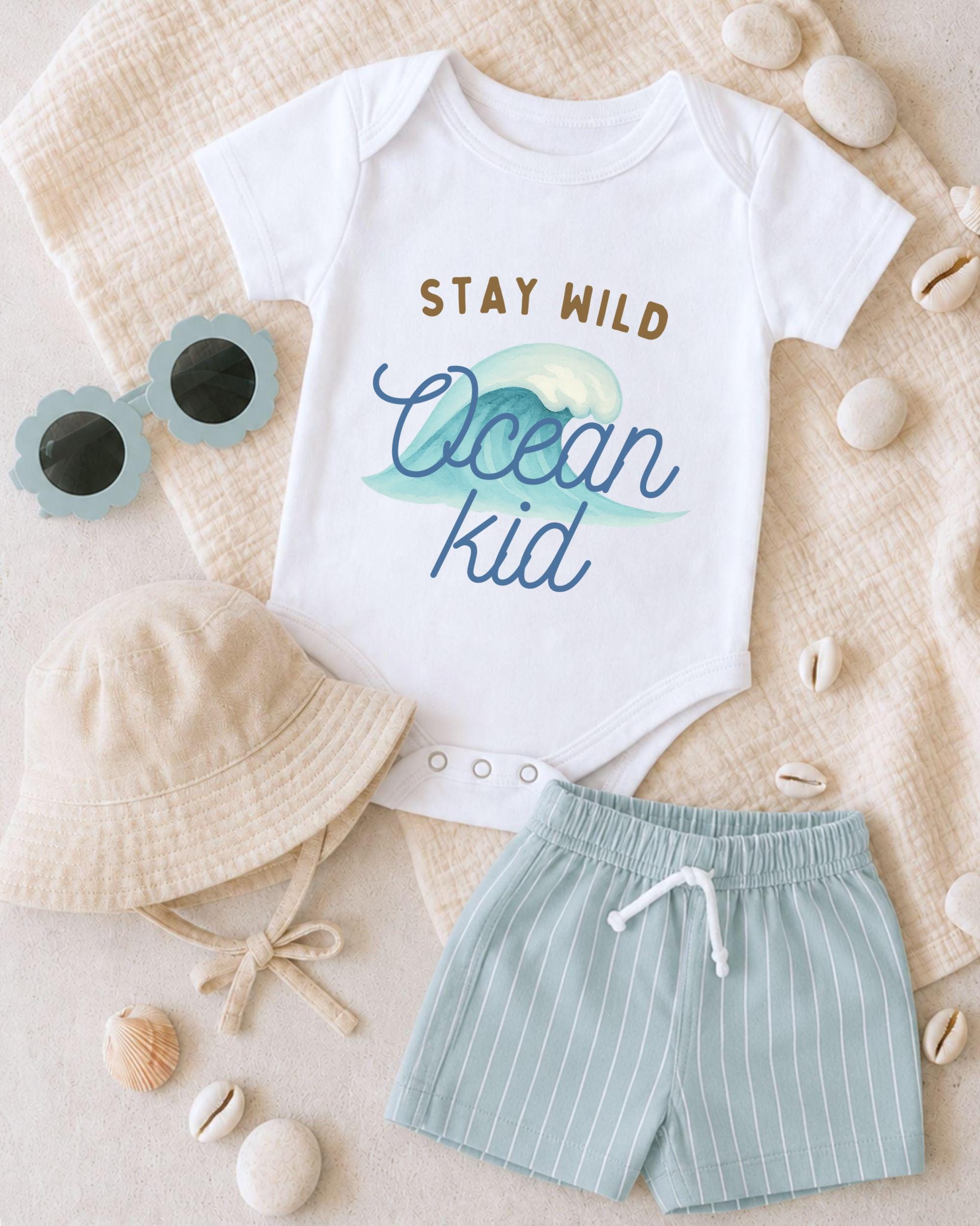 A baby's outfit, including a white onesie with the text "STAY WILD Ocean kid", a beige bucket hat, and striped shorts, along with sunglasses and seashells, are arranged on a beige surface.