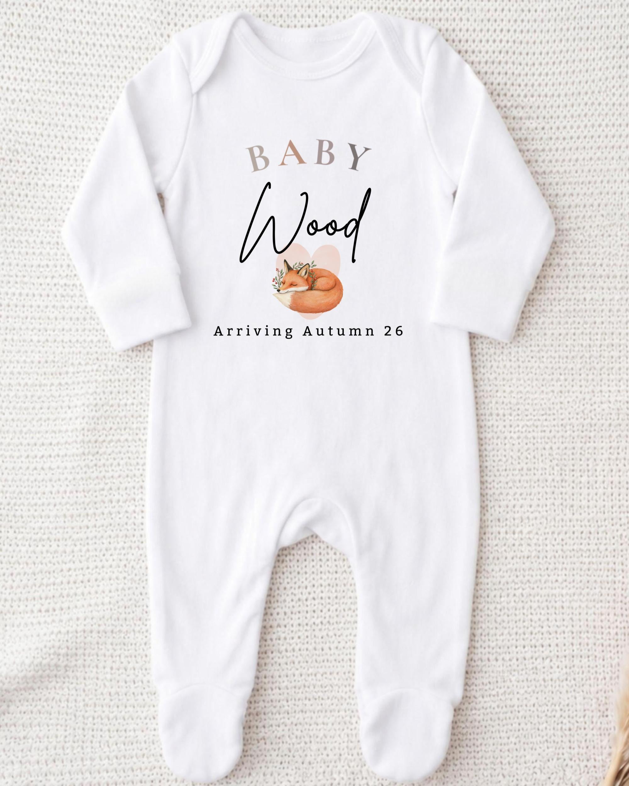 A white onesie with the text "BABY WOOD Arriving Autumn 26" printed on it.