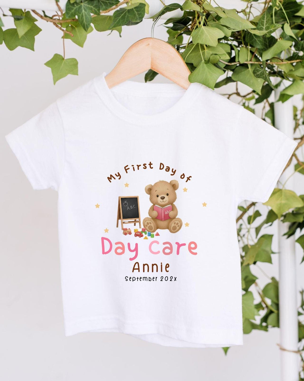A white t-shirt with a teddy bear and a chalkboard, along with the text "My First Day of Day Care Annie September 2023".