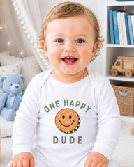 A smiling baby wearing a white onesie with a smiley face and the text "One happy dude" is sitting in a room with a teddy bear and bookshelf in the background.