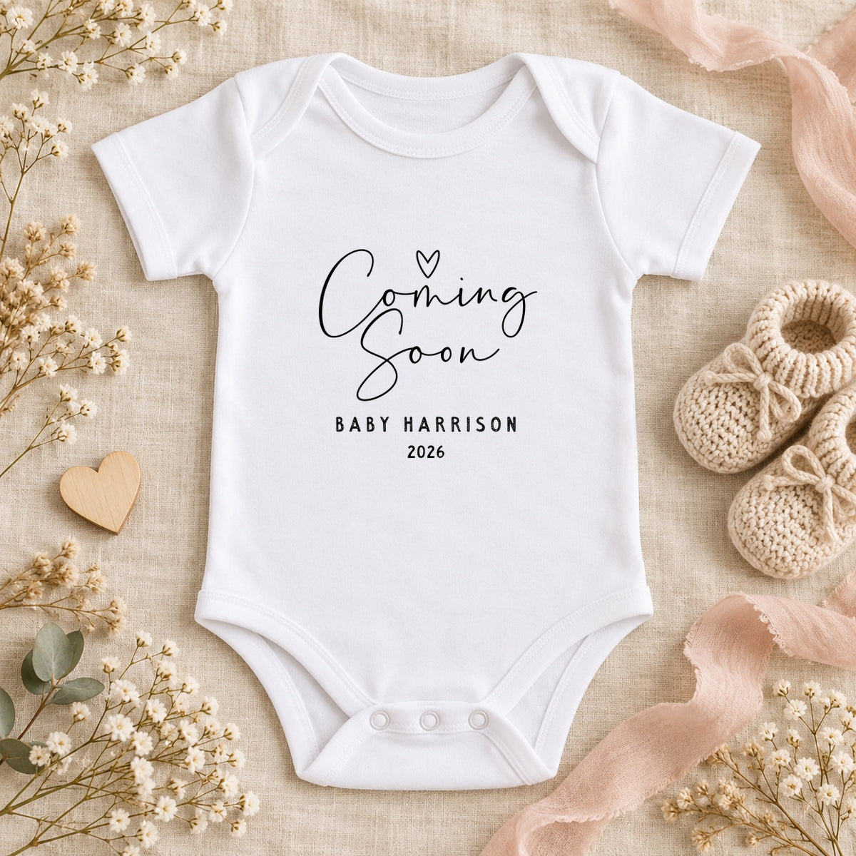 White Baby Vest with Coming Soon, Baby Harrison and 2026 in black, beside beige knitted booties, a wooden heart, flowers and blush ribbon on ivory linen.