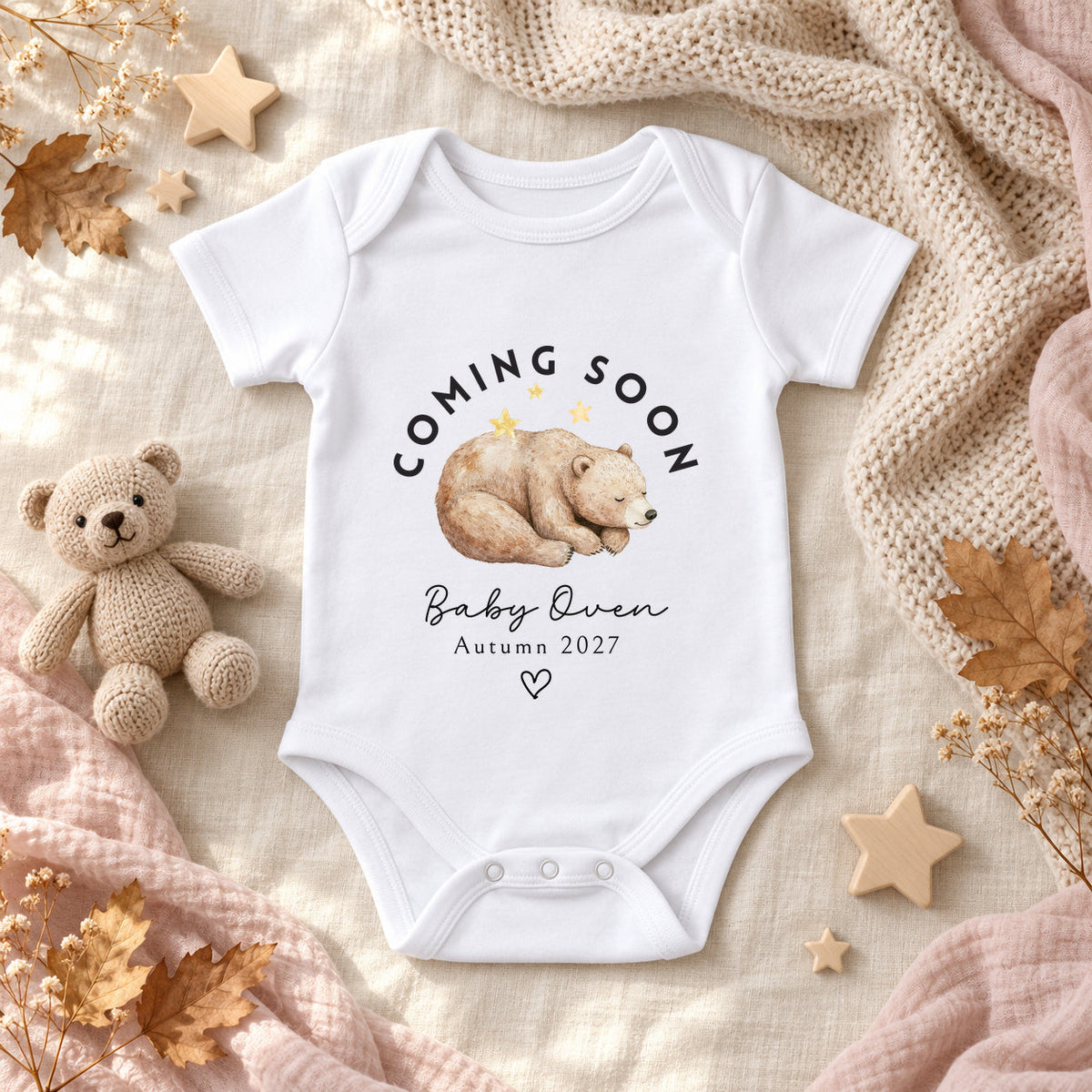 White Coming Soon bear pregnancy announcement Baby Vest personalised with Baby Owen and Autumn 2027, styled with a knitted bear, wooden stars, cream flowers, autumn leaves and blush muslin.