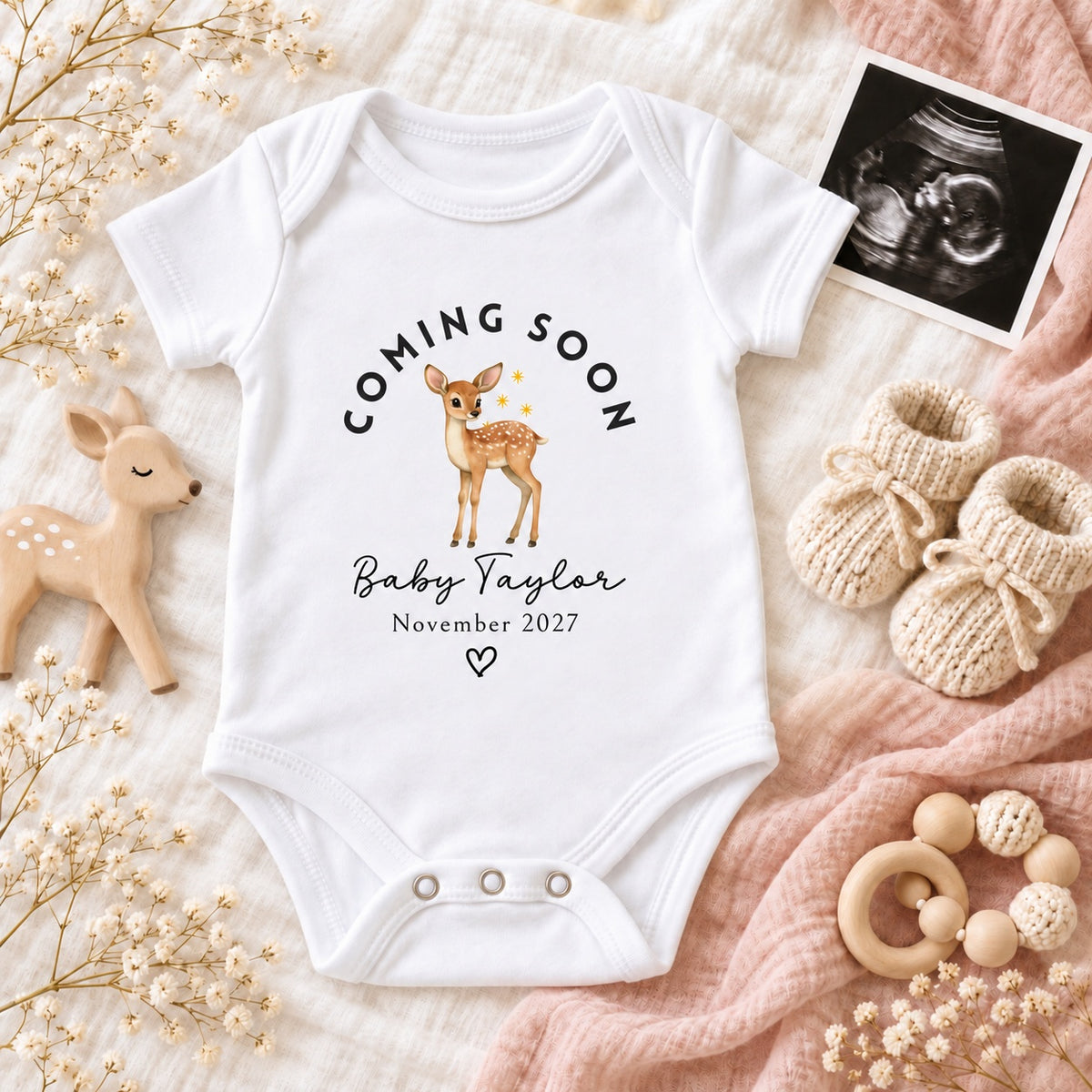 White Baby Vest printed with Coming Soon, a watercolour deer, Baby Taylor and November 2027, styled with a scan photo, wooden deer, blush muslin, knitted booties and white flowers.