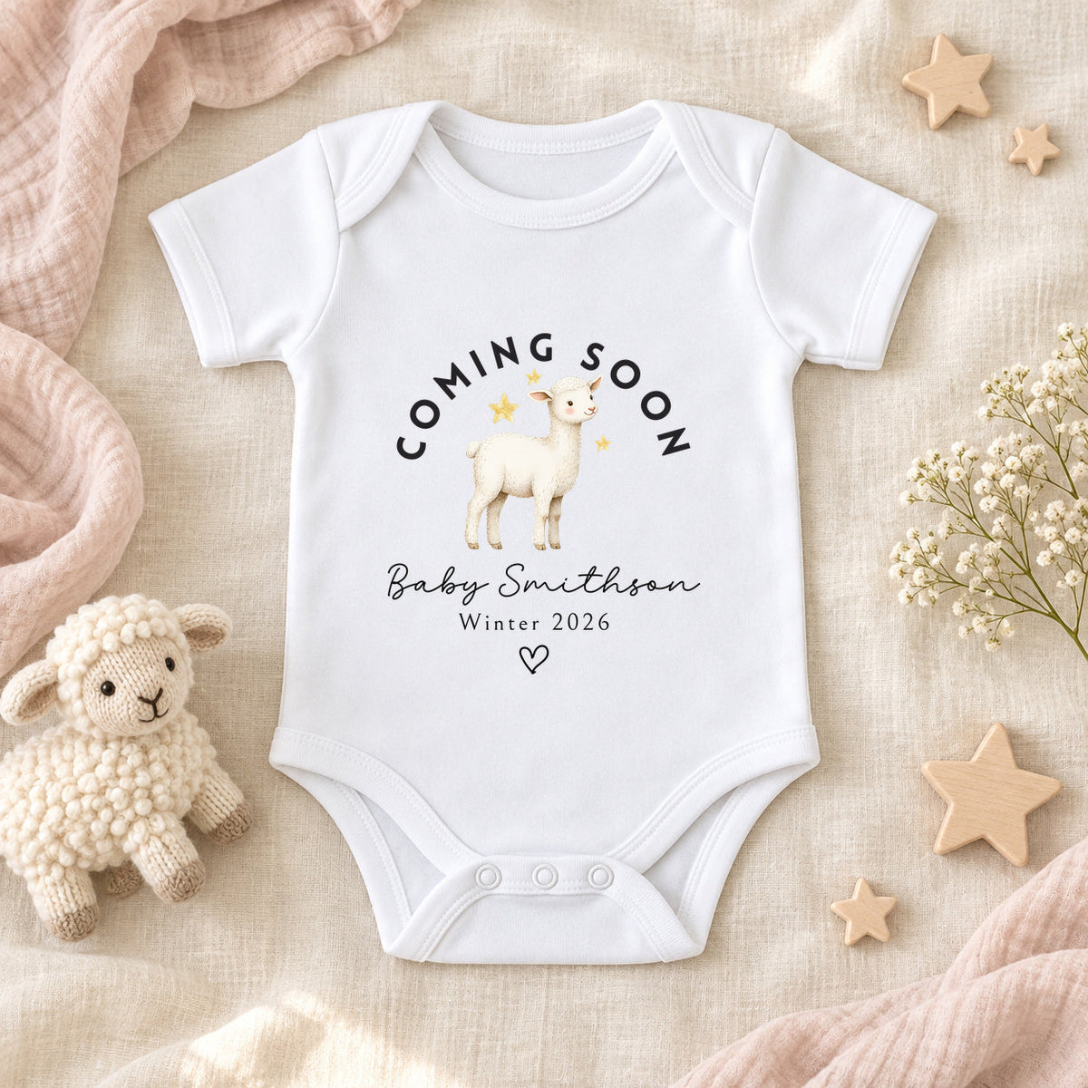 White Coming Soon lamb pregnancy announcement Baby Vest personalised with Baby Smithson and Winter 2026, styled on ivory linen with a knitted lamb, wooden stars, white flowers and blush muslin.
