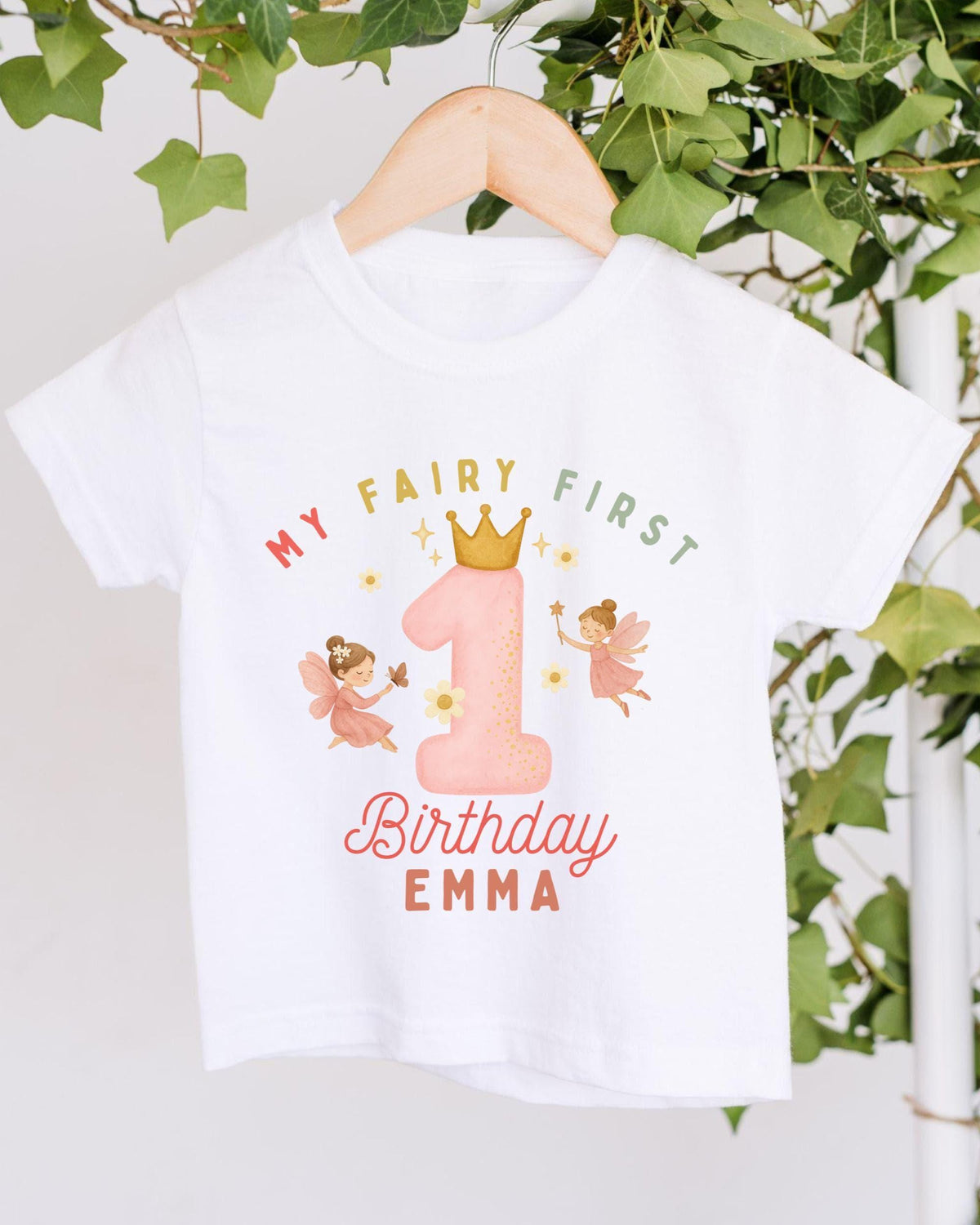 A white t-shirt with a graphic design featuring a pink number one and two-tailed fairies, along with the text "My Fairy First Birthday Emma" printed on it.