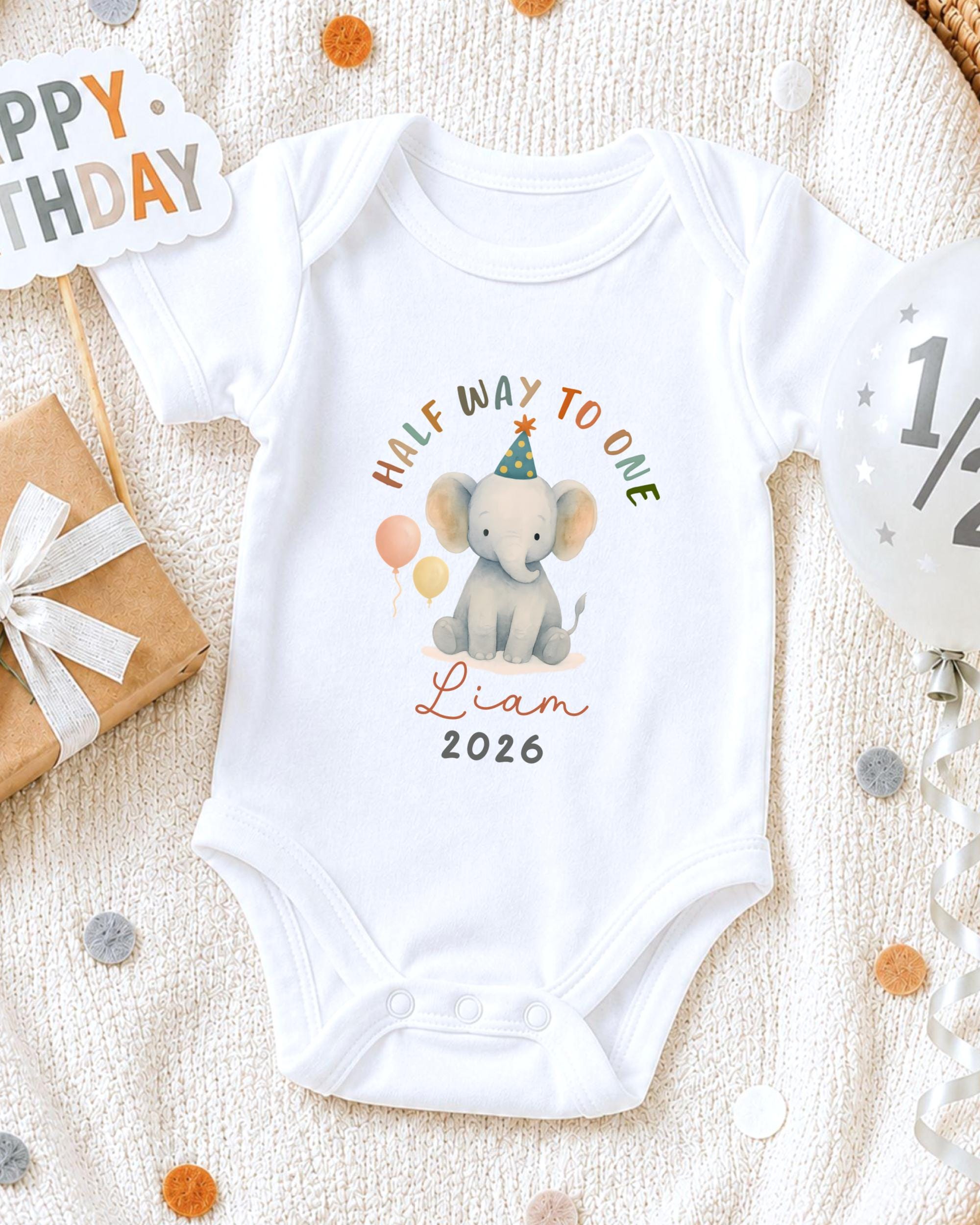 A white onesie with a cartoon elephant and the text "Half Way to One" printed on it, along with the name "Liam" and the year "2026". The onesie is placed on a white blanket, surrounded by birthday decorations such as a "Happy Birthday" sign, a balloon, and confetti.