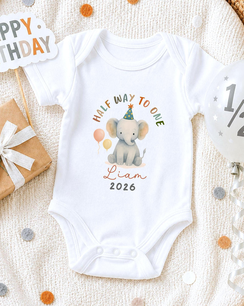 A white onesie with a cartoon elephant and the text "Half Way to One" printed on it, along with the name "Liam" and the year "2026". The onesie is placed on a white blanket, surrounded by birthday decorations such as a "Happy Birthday" sign, a balloon, and confetti.