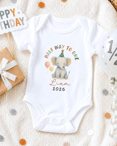 A white onesie with a cartoon elephant and the text "Half Way to One" printed on it, along with the name "Liam" and the year "2026". The onesie is placed on a white blanket, surrounded by birthday decorations such as a "Happy Birthday" sign, a balloon, and confetti.