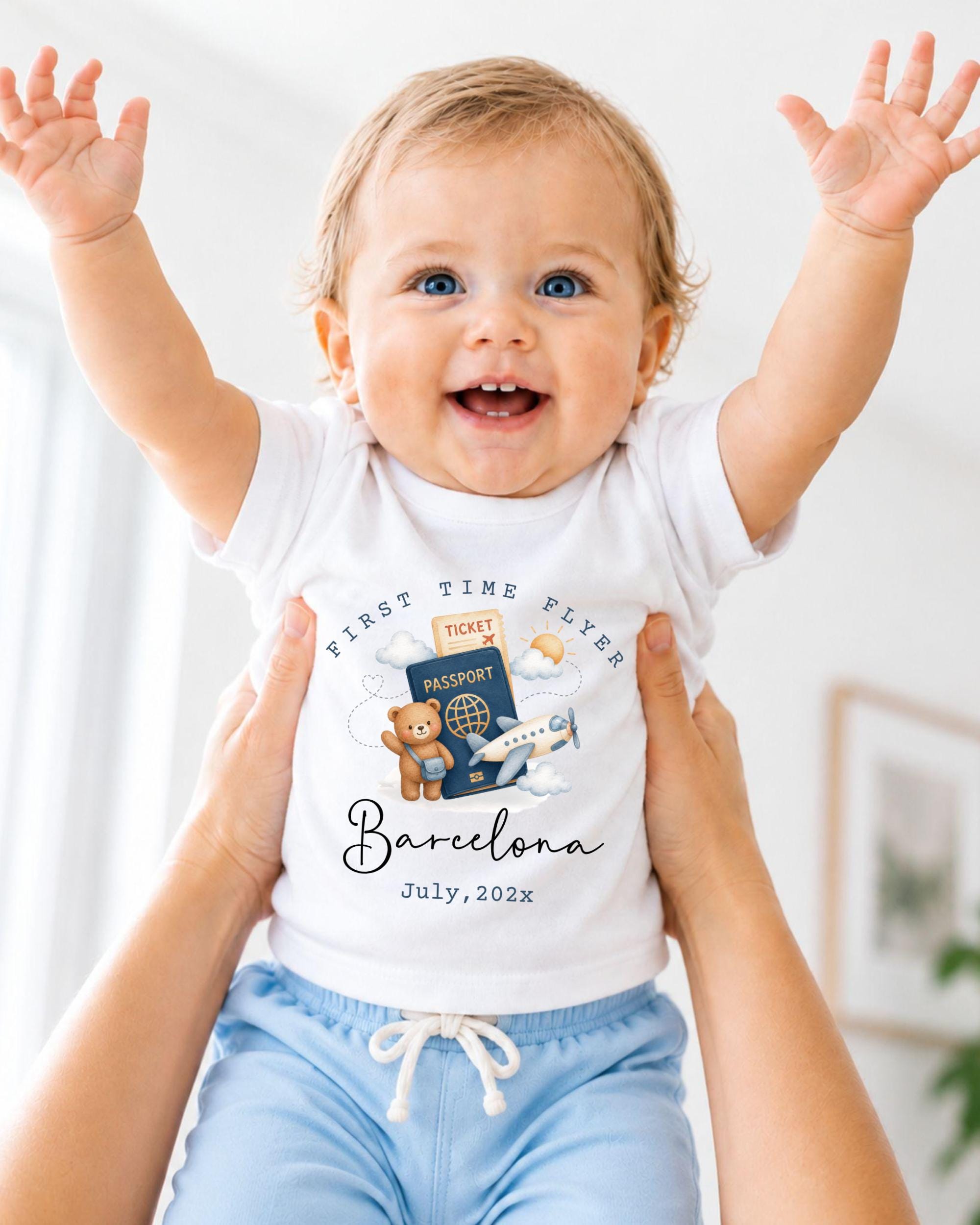 A smiling baby wearing a white t-shirt with a graphic of a passport and airplane, being held up by someone's arms.