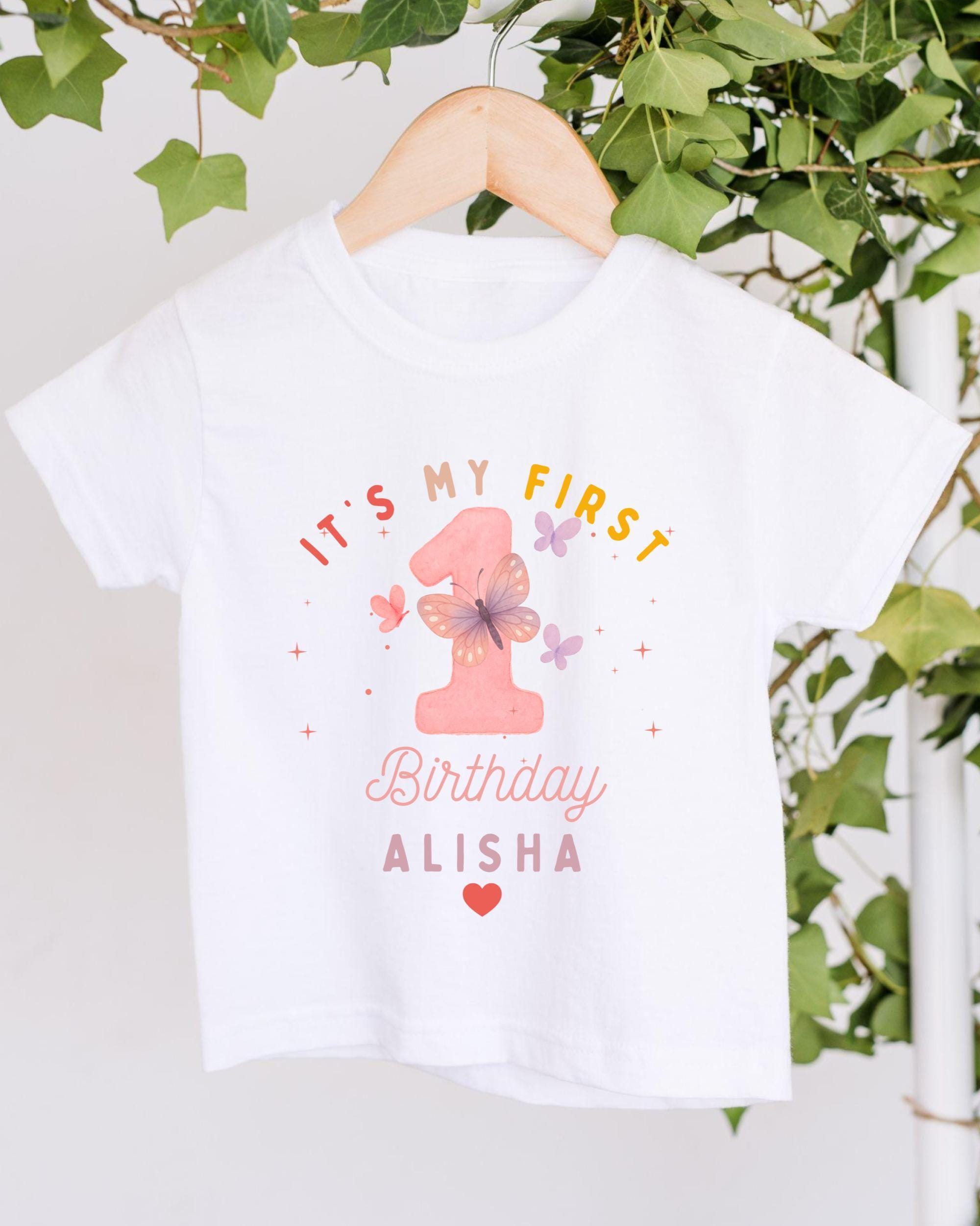 A white t-shirt with a pink number one and text that says "It's my first birthday Alisha" hanging on a wooden hanger.
