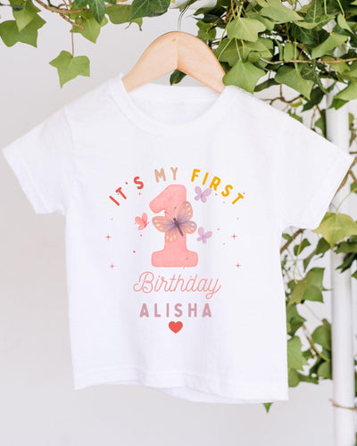 A white t-shirt with a pink number one and text that says "It's my first birthday Alisha" hanging on a wooden hanger.