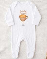 A white onesie with a graphic of a cup of coffee and the text "Locally Brewed Fredricine 2026" printed on it.