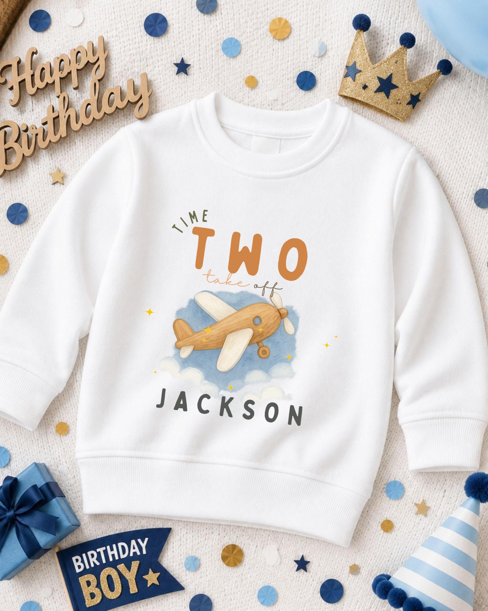 A white sweatshirt with a cartoon airplane design and the text "I'm the two take off Jackson" printed on it, surrounded by birthday decorations and a crown.