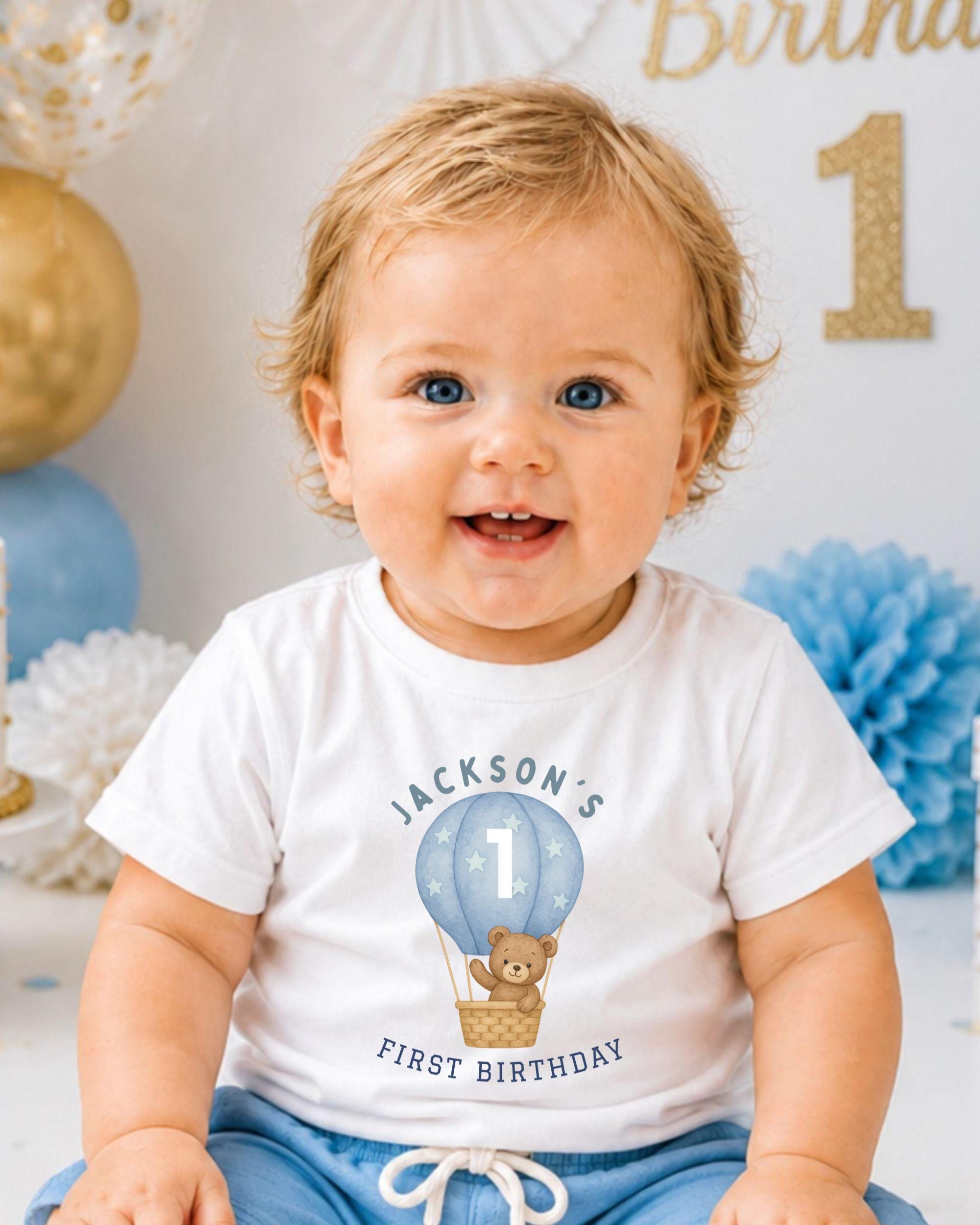 A smiling baby boy wearing a white t-shirt with a cartoon image of a hot air balloon and the text "Jackson's 1st Birthday" printed on it.