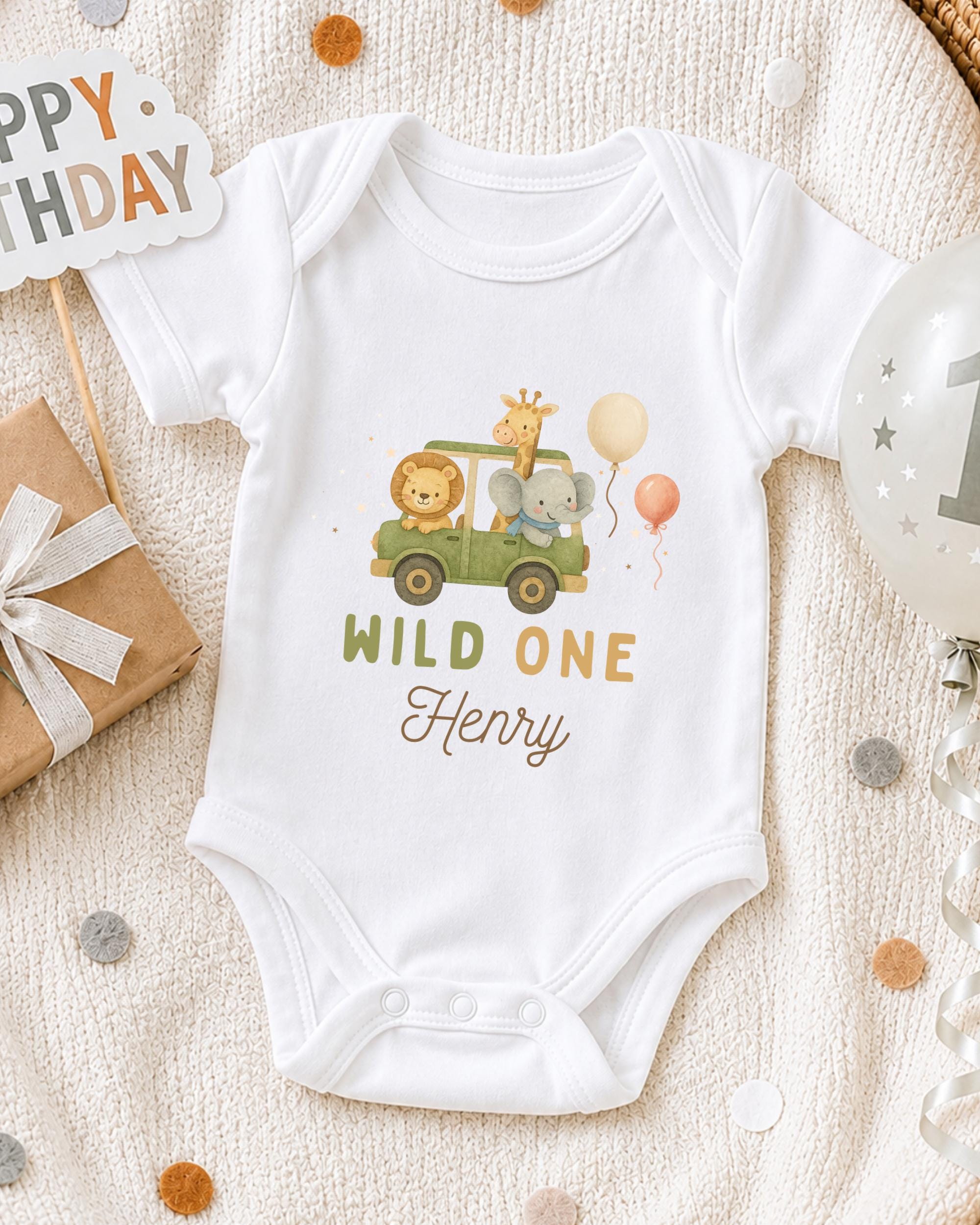 A white onesie with a cartoon image of a giraffe and elephant, along with the text "Wild One Henry", is displayed on a bed with a gift box and a birthday balloon.