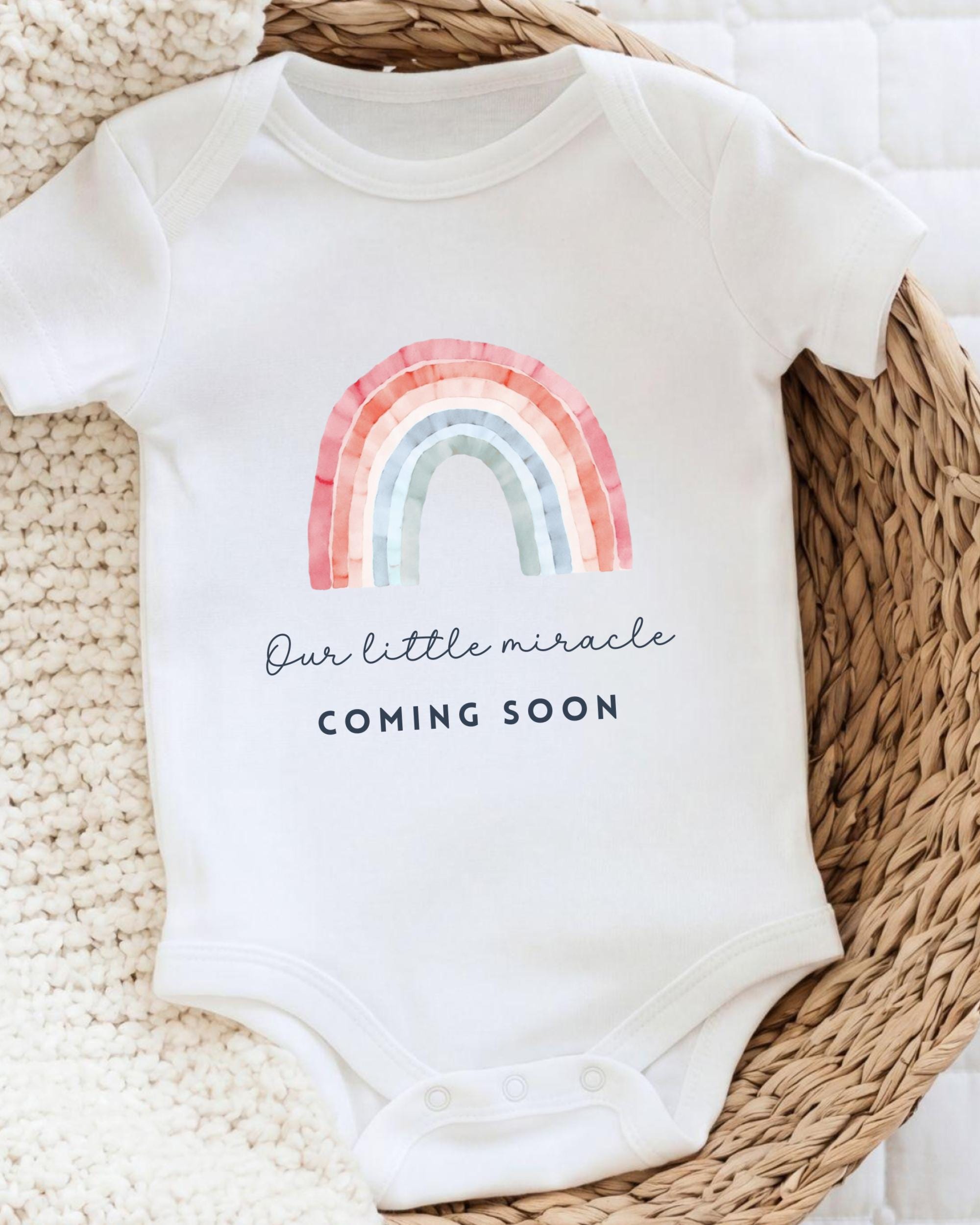 A white baby onesie with a rainbow graphic and the text "Our little miracle coming soon" printed on it.
