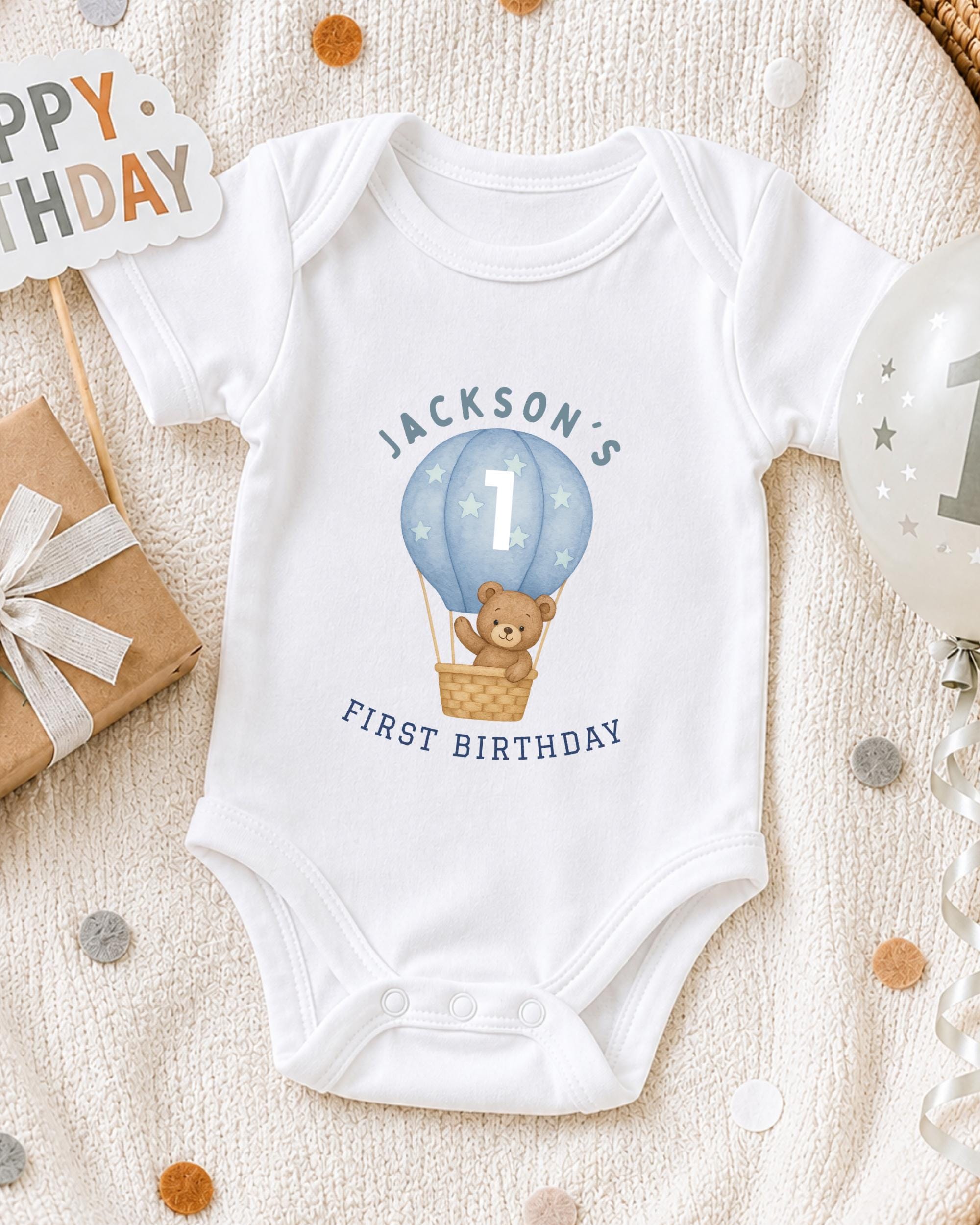 A white onesie with a teddy bear and a hot air balloon graphic, along with a "Happy Birthday Jackson's First Birthday" sign, is displayed on a bed with confetti.