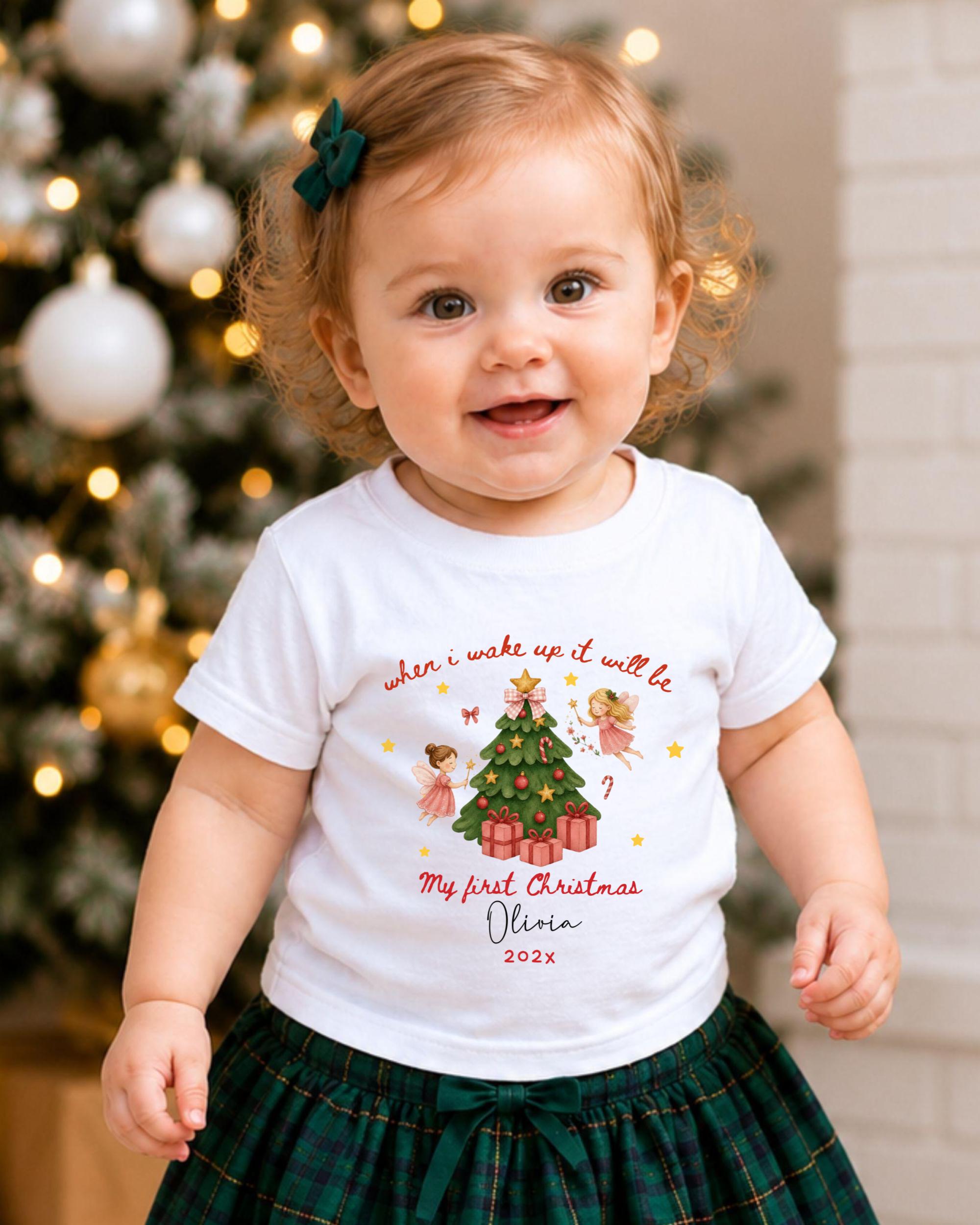 A smiling baby girl wearing a white t-shirt with a Christmas tree design and text, standing in front of a Christmas tree with lights.