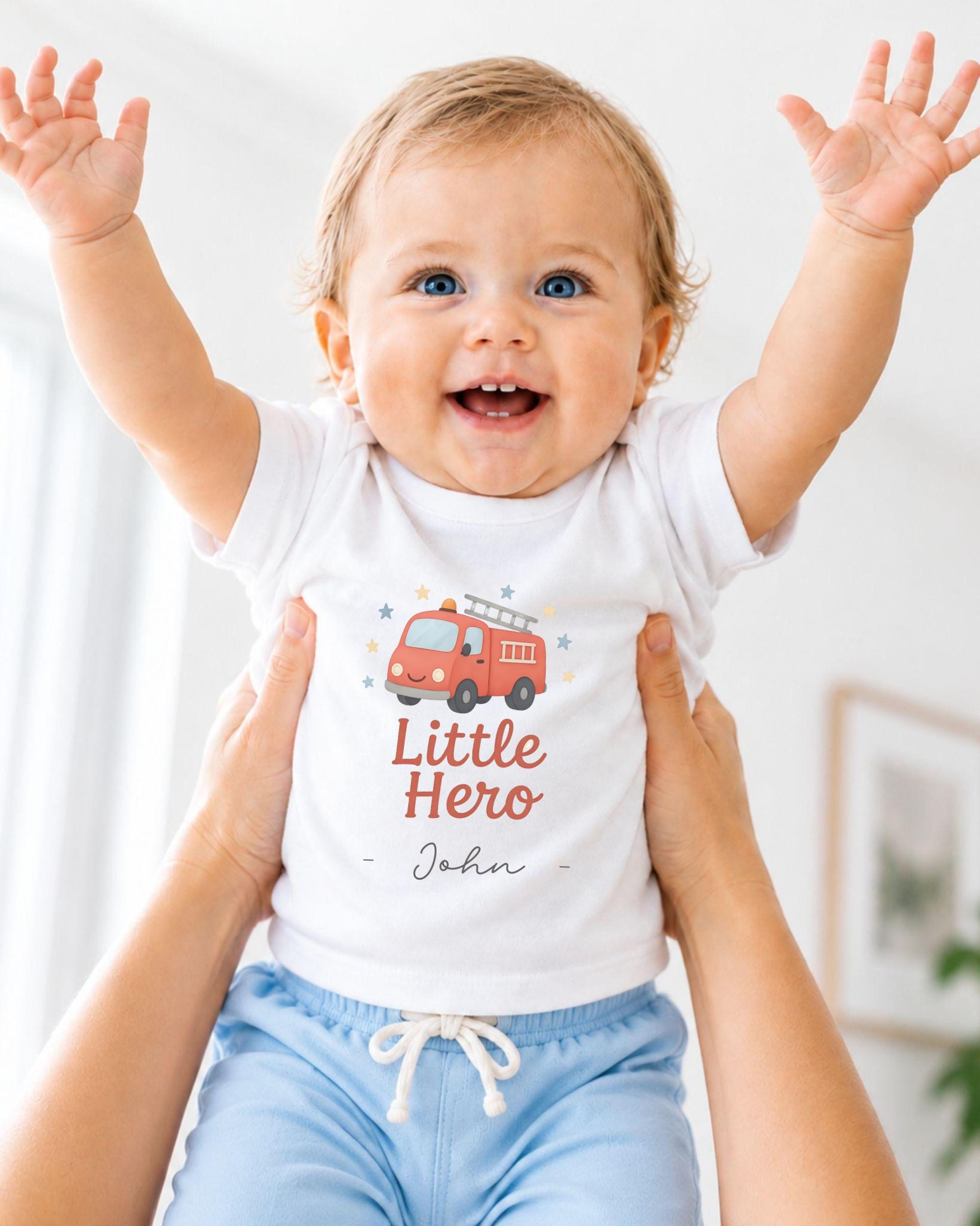 A smiling baby wearing a white t-shirt with a fire truck graphic and the text "Little Hero" is being held up by someone's arms.