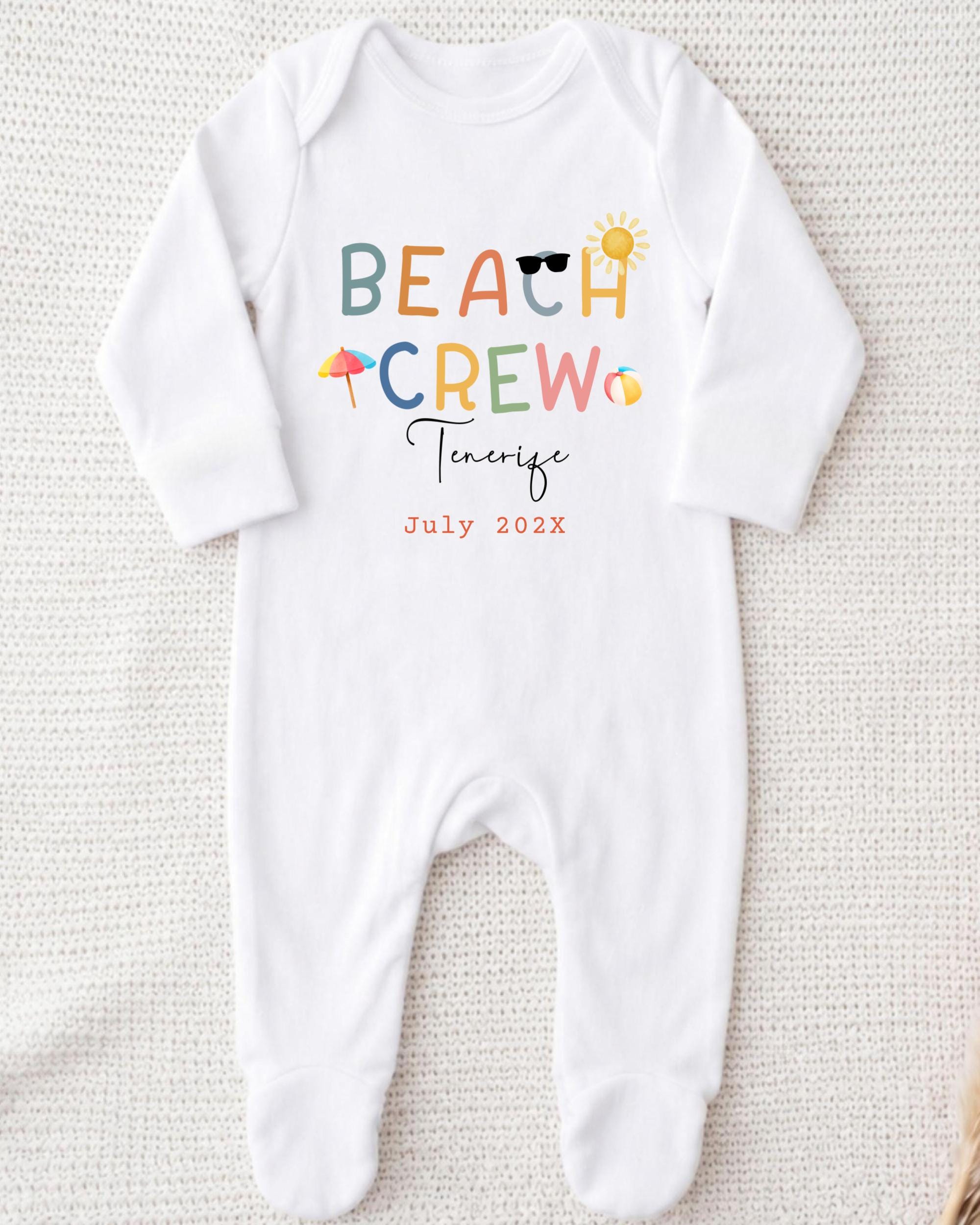 A white onesie with the text "BEACH CREW" printed on it, along with various beach-related icons such as a sun, sunglasses, and an umbrella.