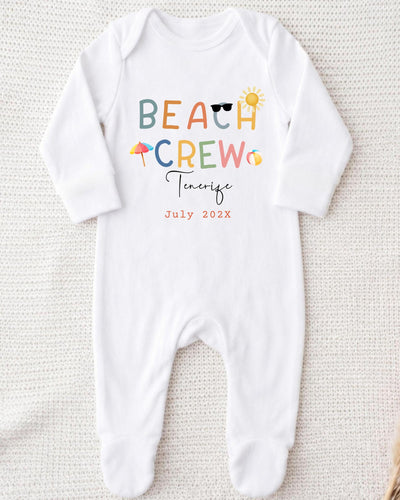 A white onesie with the text "BEACH CREW" printed on it, along with various beach-related icons such as a sun, sunglasses, and an umbrella.