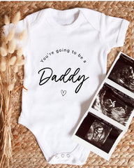 A white baby onesie with the text "You're going to be a Daddy" printed on it, along with a heart symbol. The onesie is placed next to a set of three black and white images depicting an ultrasound image of a baby.