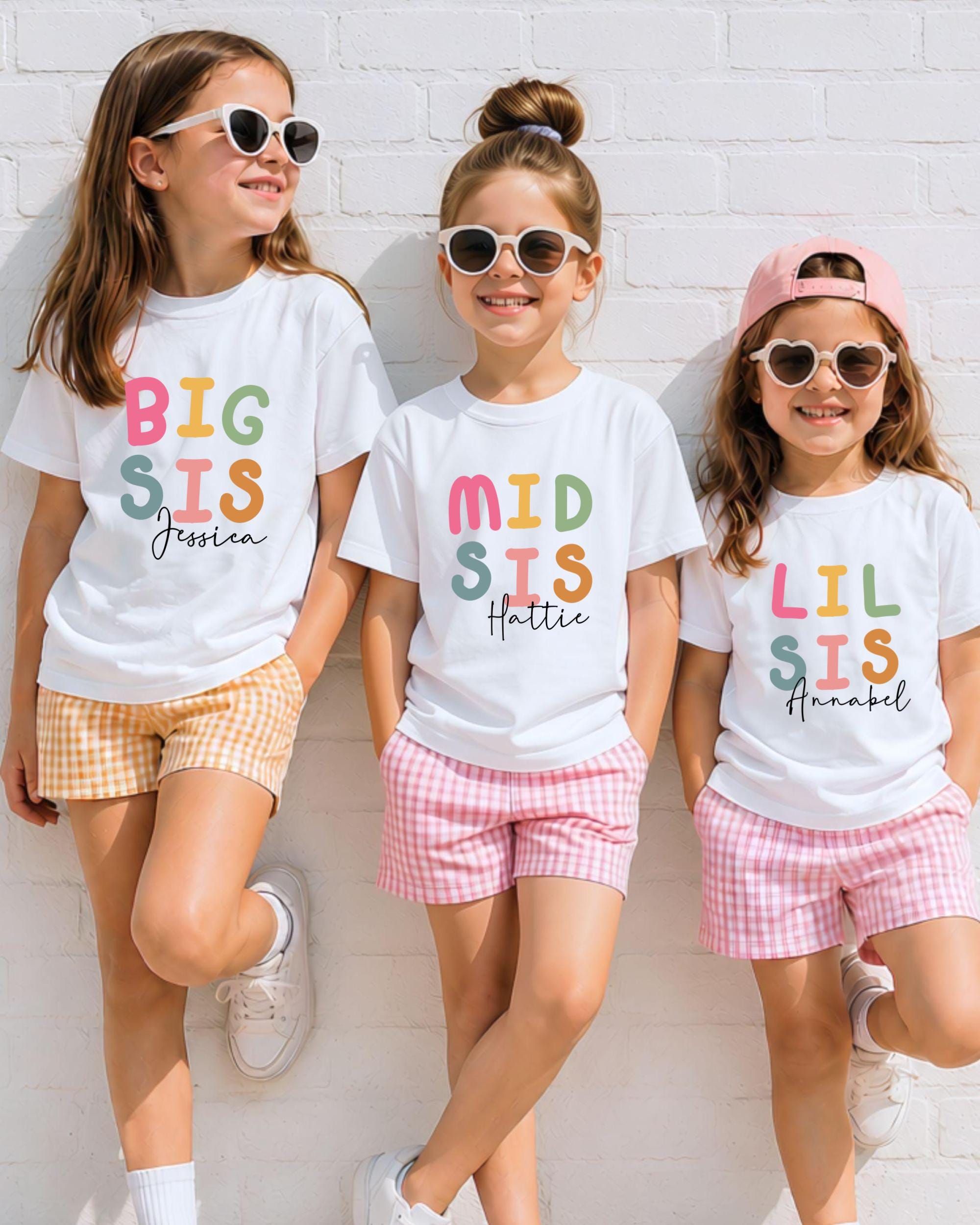 Three young girls wearing white t-shirts with colorful text and sunglasses, standing against a white brick wall.