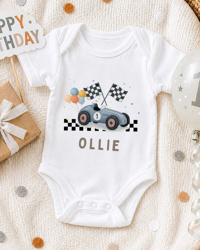 A white bodysuit with a race car design and checkered flags, along with a birthday sign and confetti, is displayed on a bed.