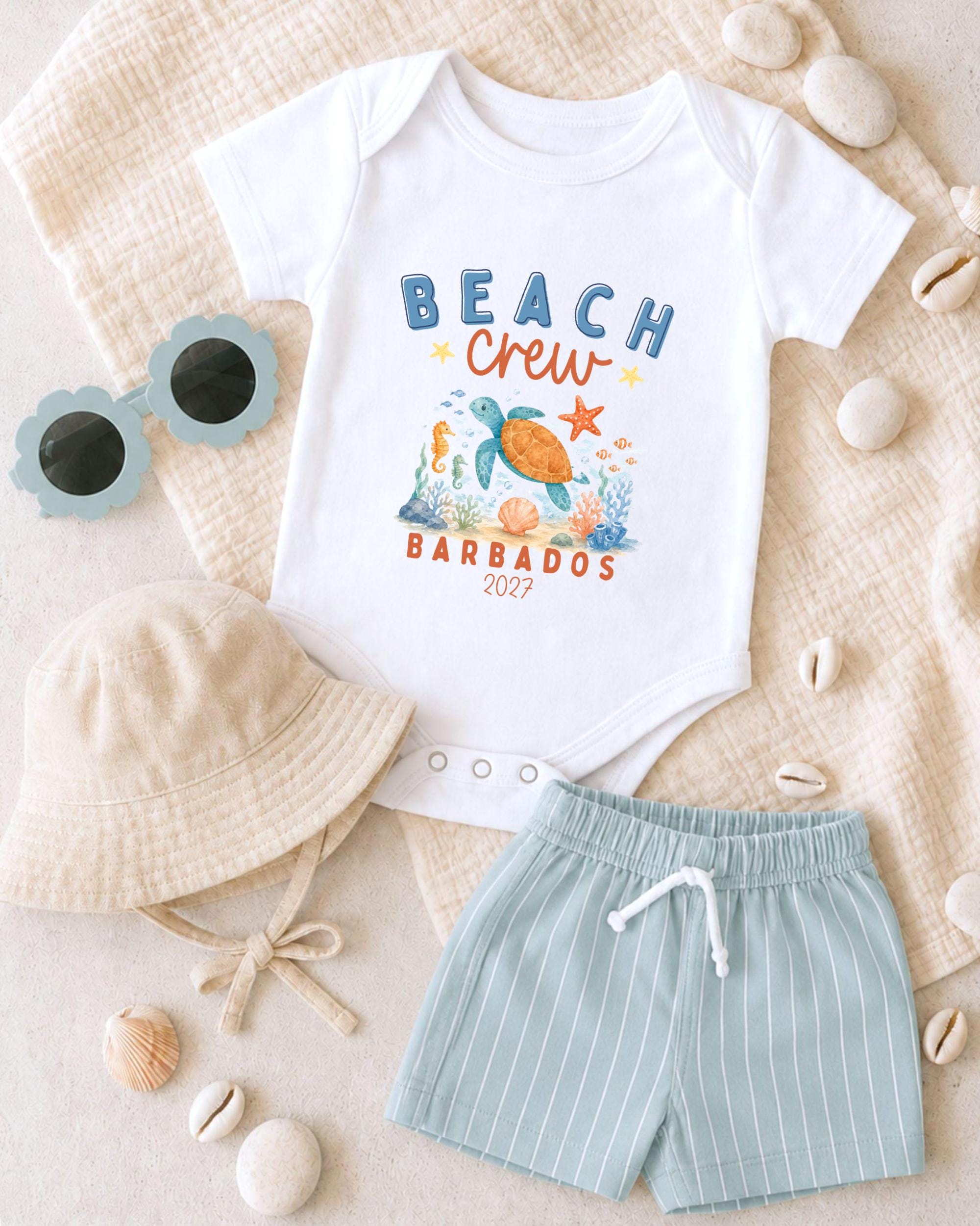 A baby's outfit, including a white onesie with a beach crew design, striped shorts, and a beige bucket hat, along with sunglasses and seashells, is laid out on a beige surface.