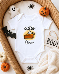 A white onesie with a pumpkin and ghost design, along with Halloween decorations, is displayed in a basket.