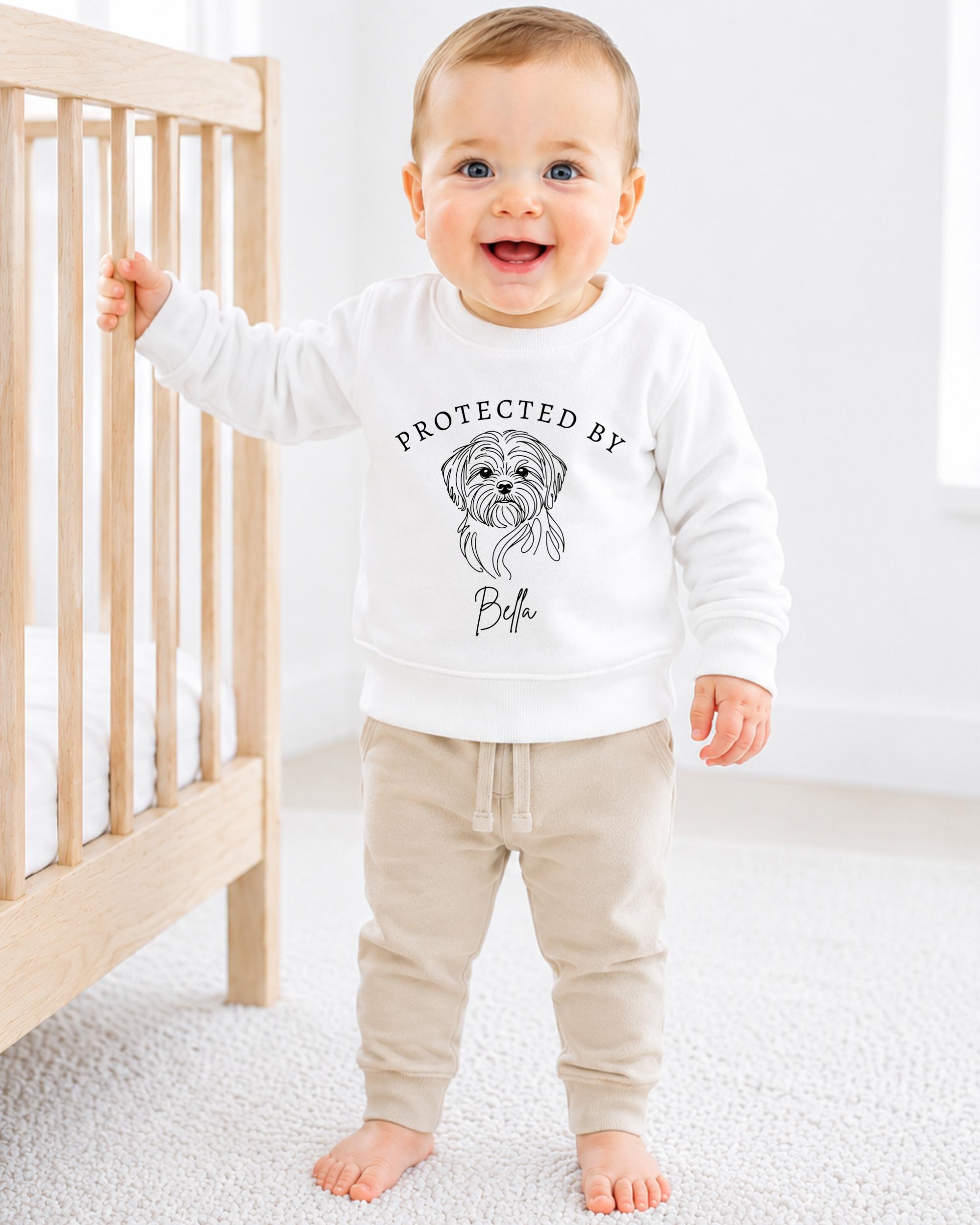 A smiling baby boy wearing a white sweatshirt with a graphic of a dog and the text "PROTECTED BY" printed on it.