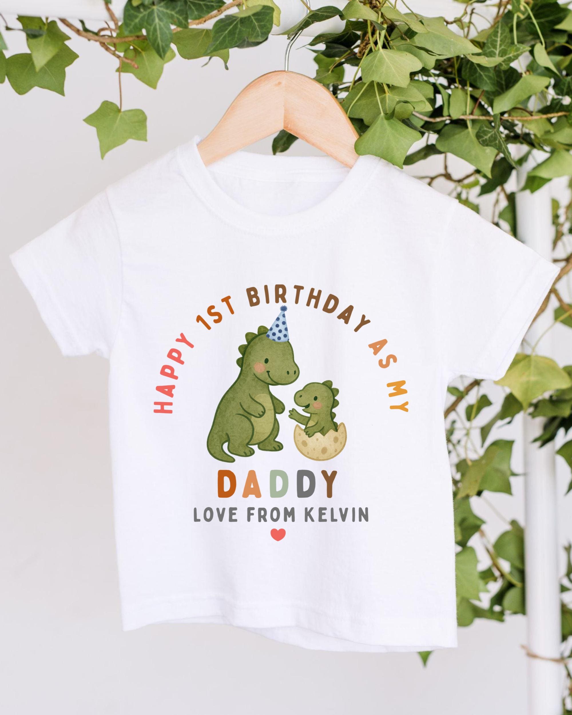A white t-shirt with a graphic design featuring a dinosaur and a baby dinosaur, along with text that reads "Happy 1st Birthday as my Daddy Love from Kevin".