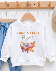 A white sweatshirt with a pink airplane graphic and the text "Noah's first flight" printed on it, hanging on a wooden hanger.