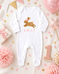 A white onesie with a bunny design and the name "EMILY" printed on it, along with a pink and gold birthday decoration, a pink star, and a pink and gold confetti arrangement.