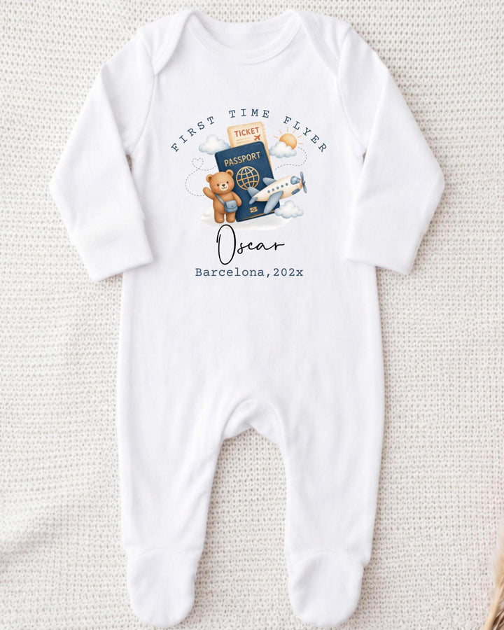 A white onesie with a graphic design depicting a teddy bear, a passport, and an airplane, along with the text "First Time Flyer Oscar Barcelona, 2023".