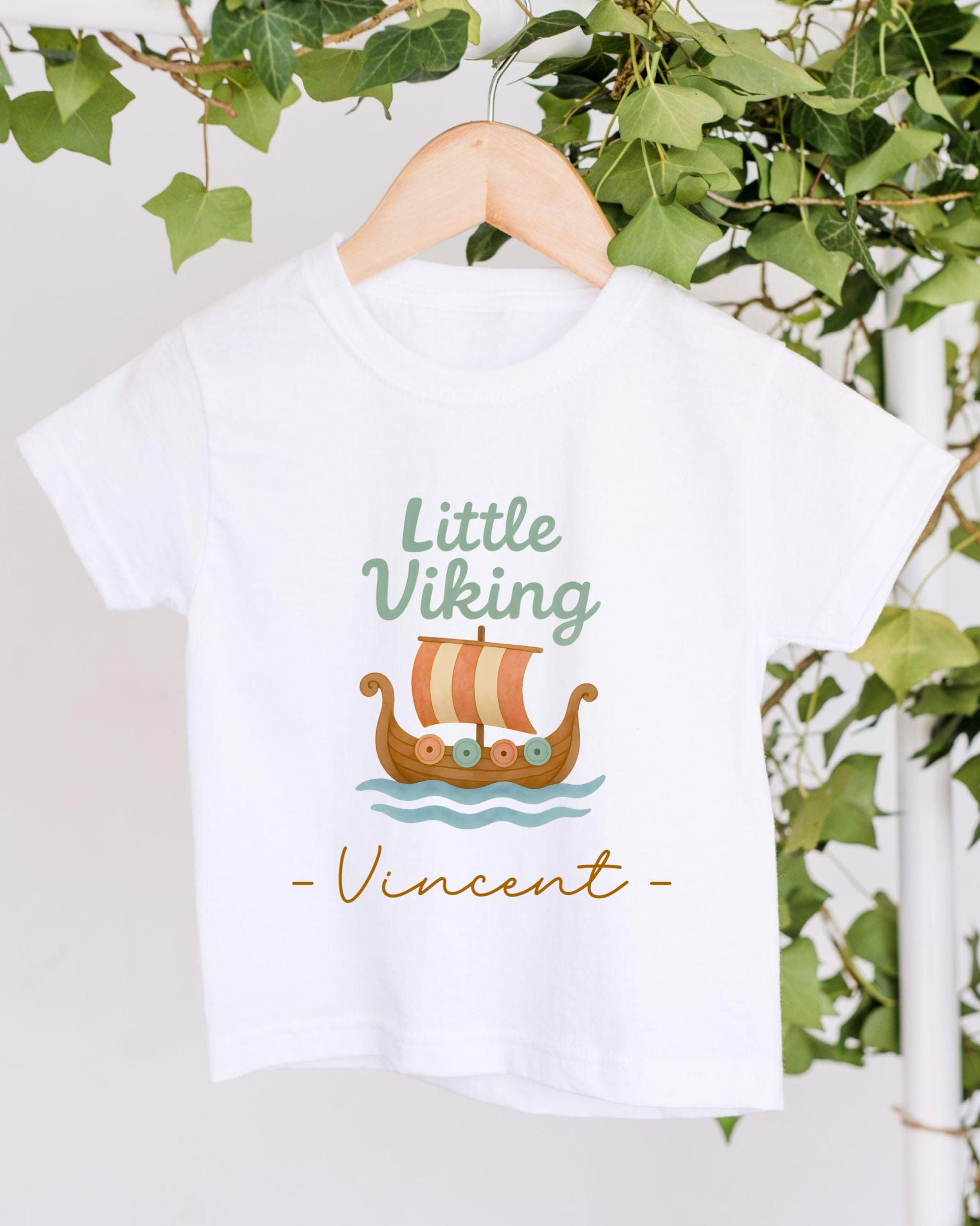 A white t-shirt with a graphic design of a Viking ship and the text "Little Viking Vincent" printed on it, hanging on a wooden hanger.