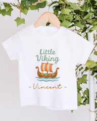 A white t-shirt with a graphic design of a Viking ship and the text "Little Viking Vincent" printed on it, hanging on a wooden hanger.
