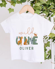 A white t-shirt with a graphic design featuring a giraffe, elephant, and other animals, along with the text "wild one Oliver".
