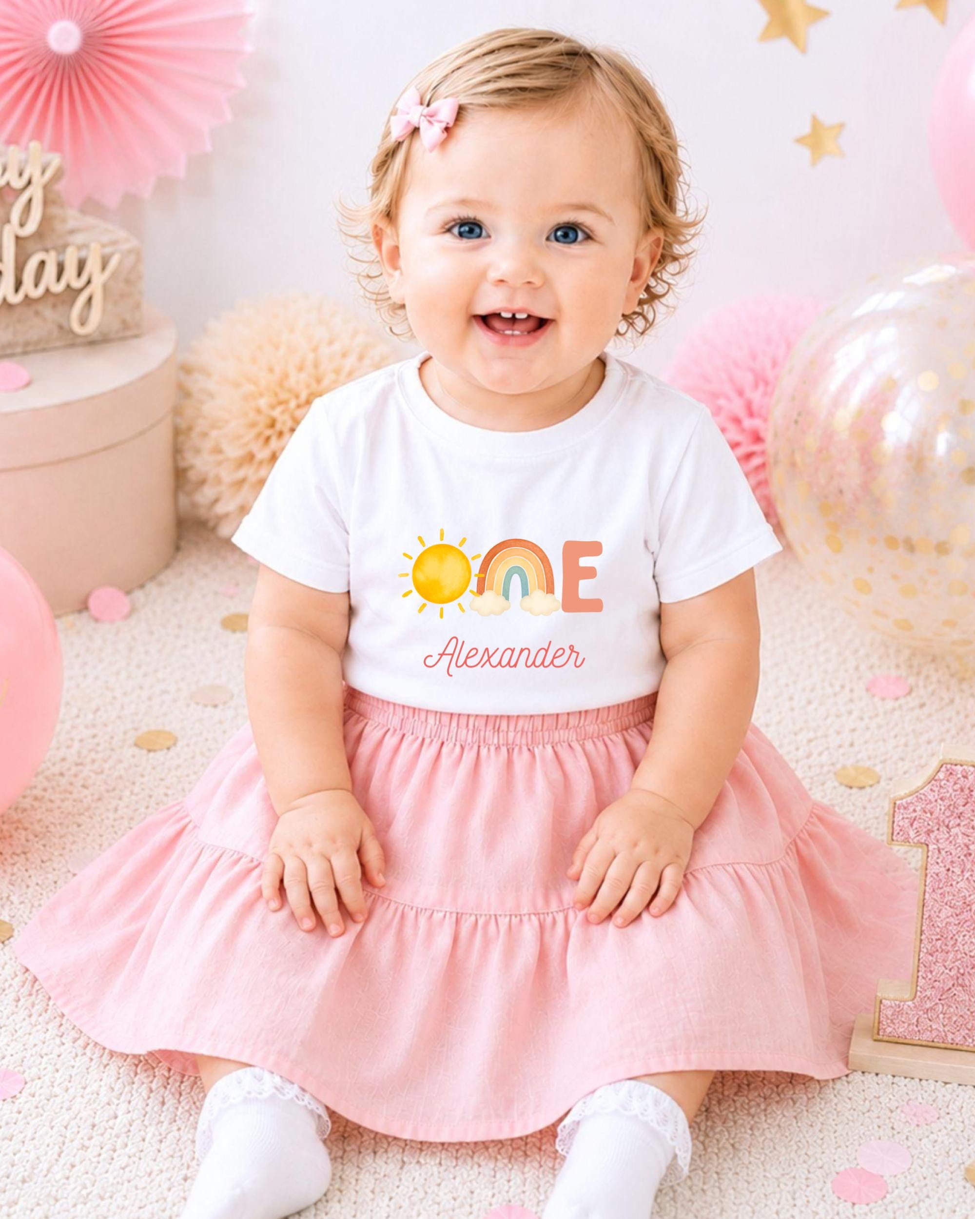 A smiling baby girl wearing a white t-shirt with a rainbow graphic and the name "Alexander" on it, sitting on a pink and white background with balloons and confetti.